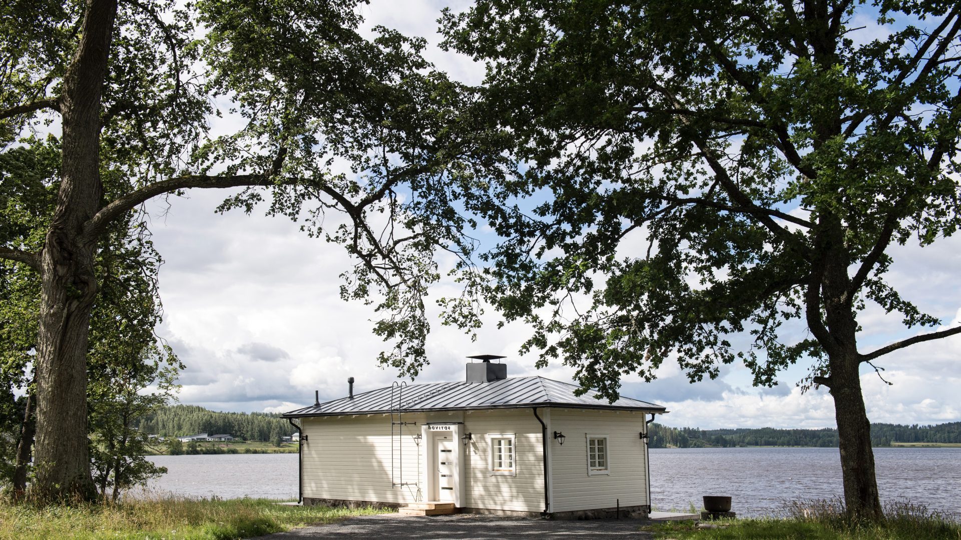 Valkoinen kesämökki vesistön äärellä suurten puiden suojassa.