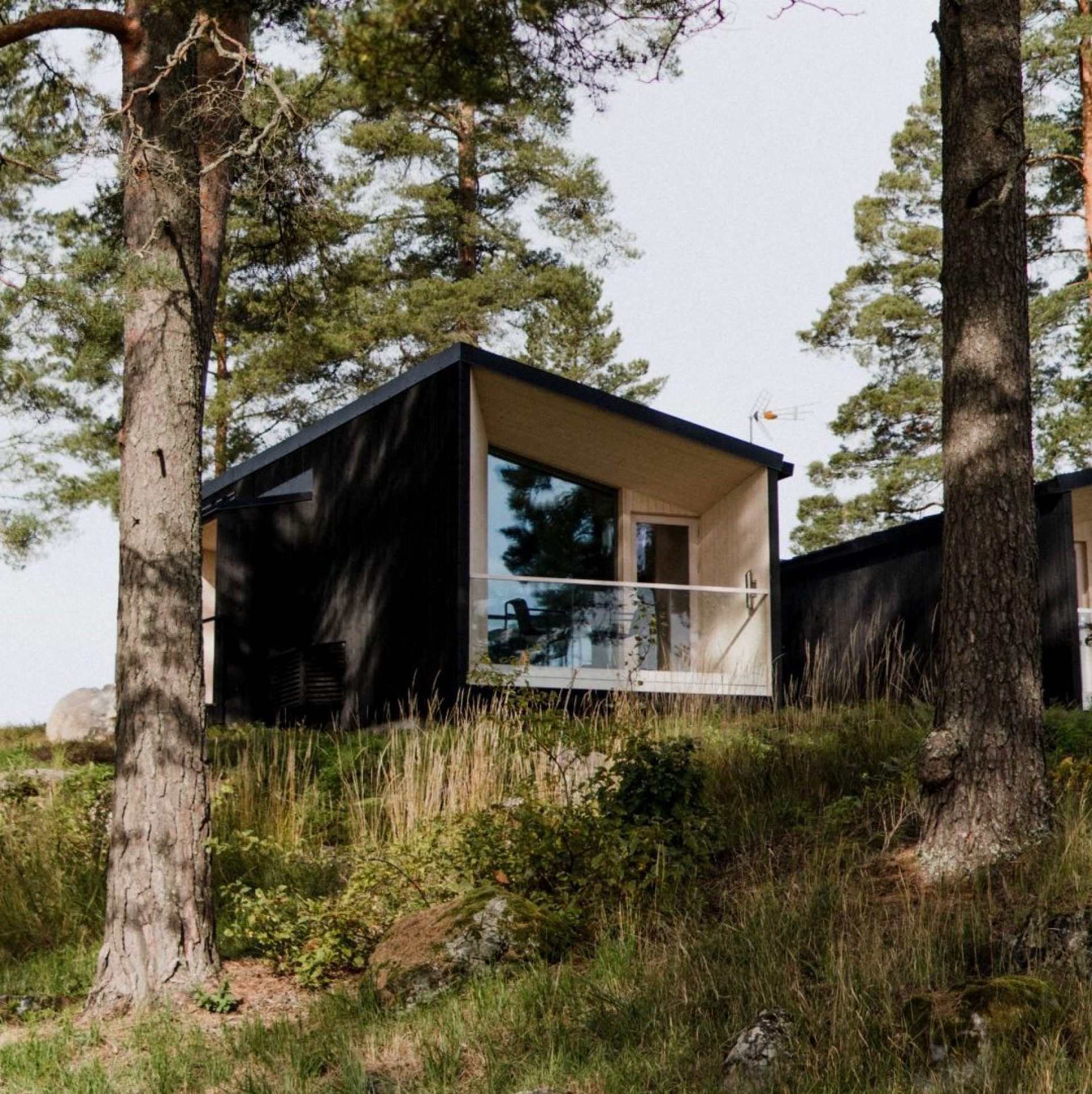Moderni mökki, jossa on lasiseinä ja parveke, sijaitsee metsäisessä ympäristössä.