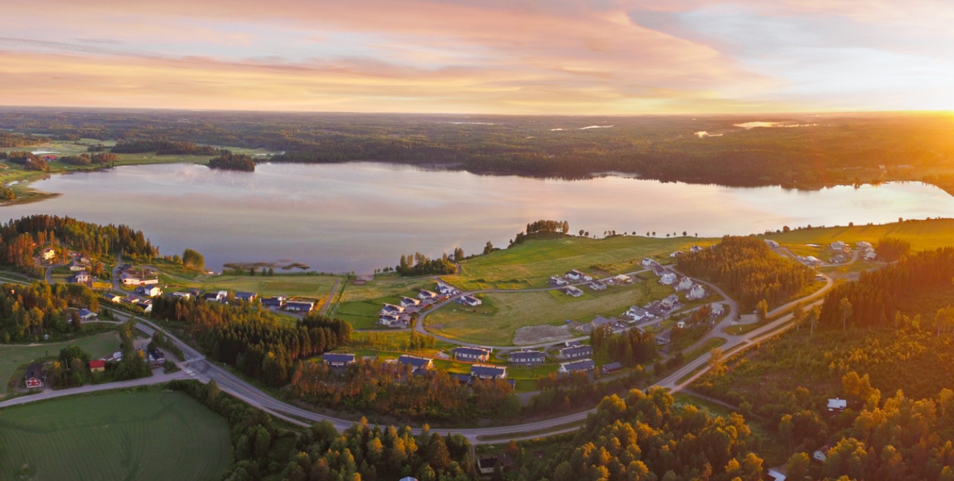 Auringonlasku hehkuu järven yllä, ympäröivien peltojen ja metsien luodessa rauhallisen maaseutumaiseman.