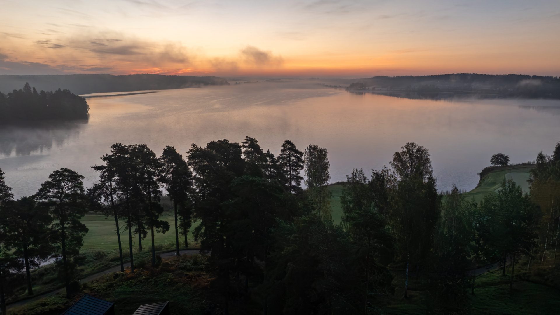 Aamuauringon ensisäteet heijastuvat rauhalliseen järveen, jota ympäröivät metsät ja sumu.