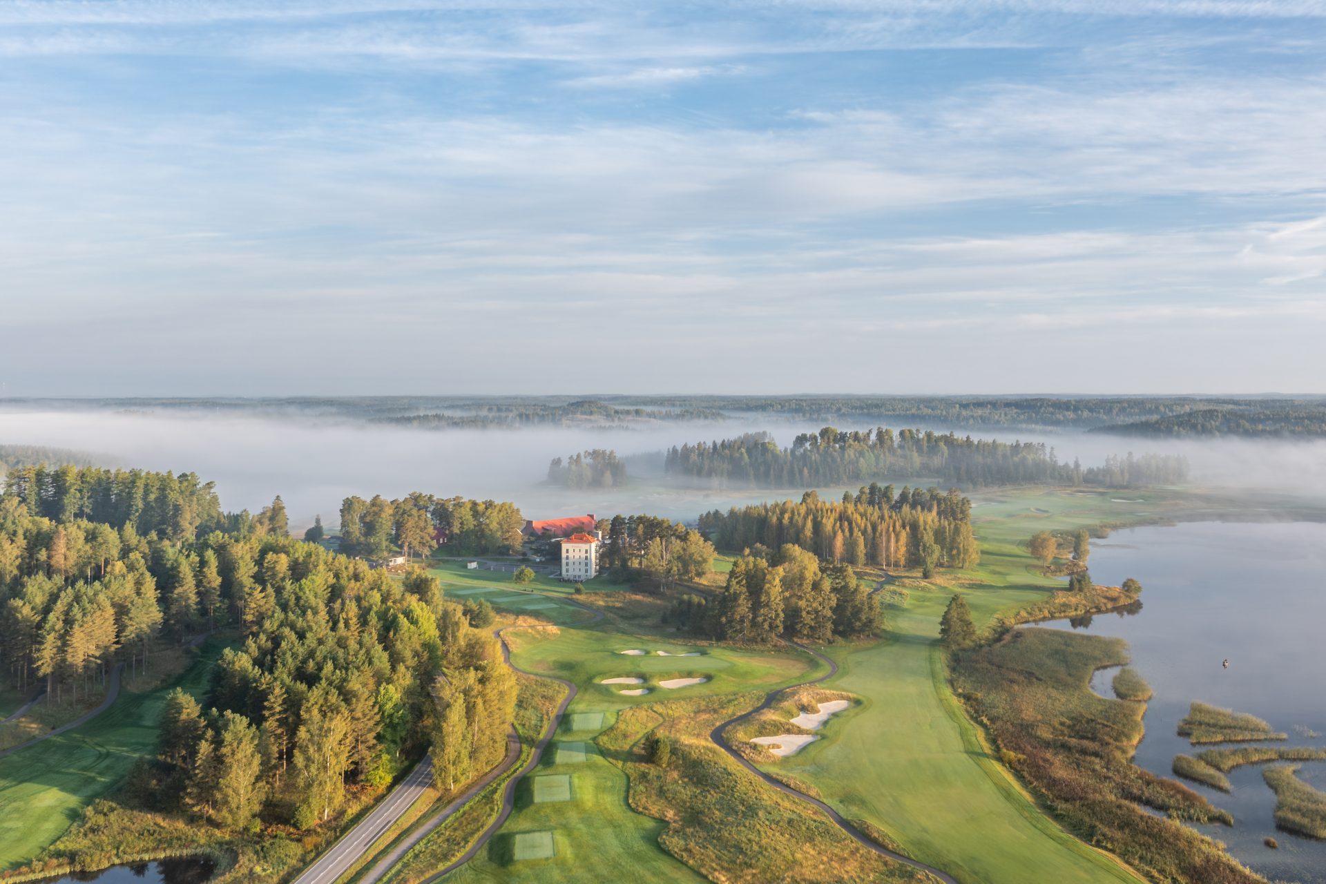 Kaunis golfkenttä ja historiallinen rakennus järven rannalla, ympärillä sumua ja vehreitä metsiä.
