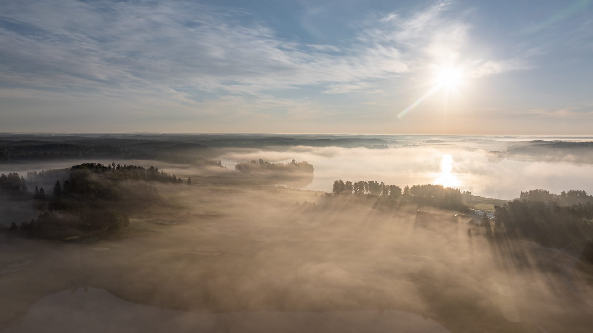Auringonlasku sumuisen golfkentän ylle järven äärellä, metsien ja sumun peittämä maisema.
